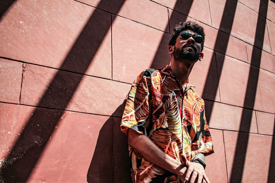 A man wearing a vibrant tropical shirt poses confidently against a wall, casting strong shadows.