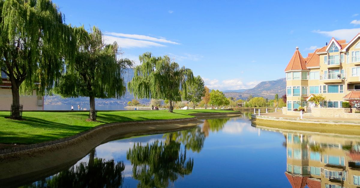 Beautiful lakefront view with reflections, trees, and a luxury building in a sunny setting.