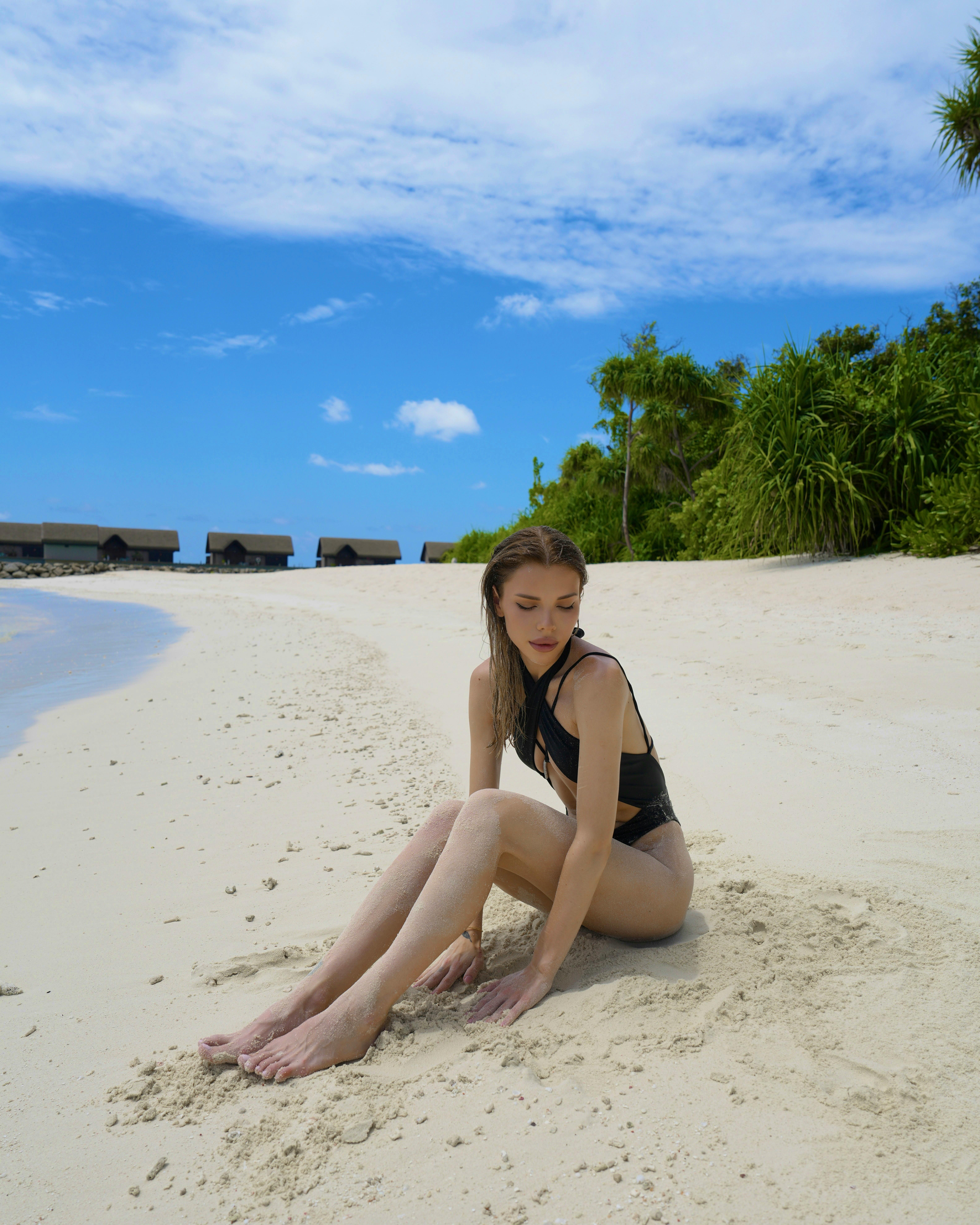 A woman in a bikini sitting on a tropical beach, surrounded by lush greenery and clear skies.