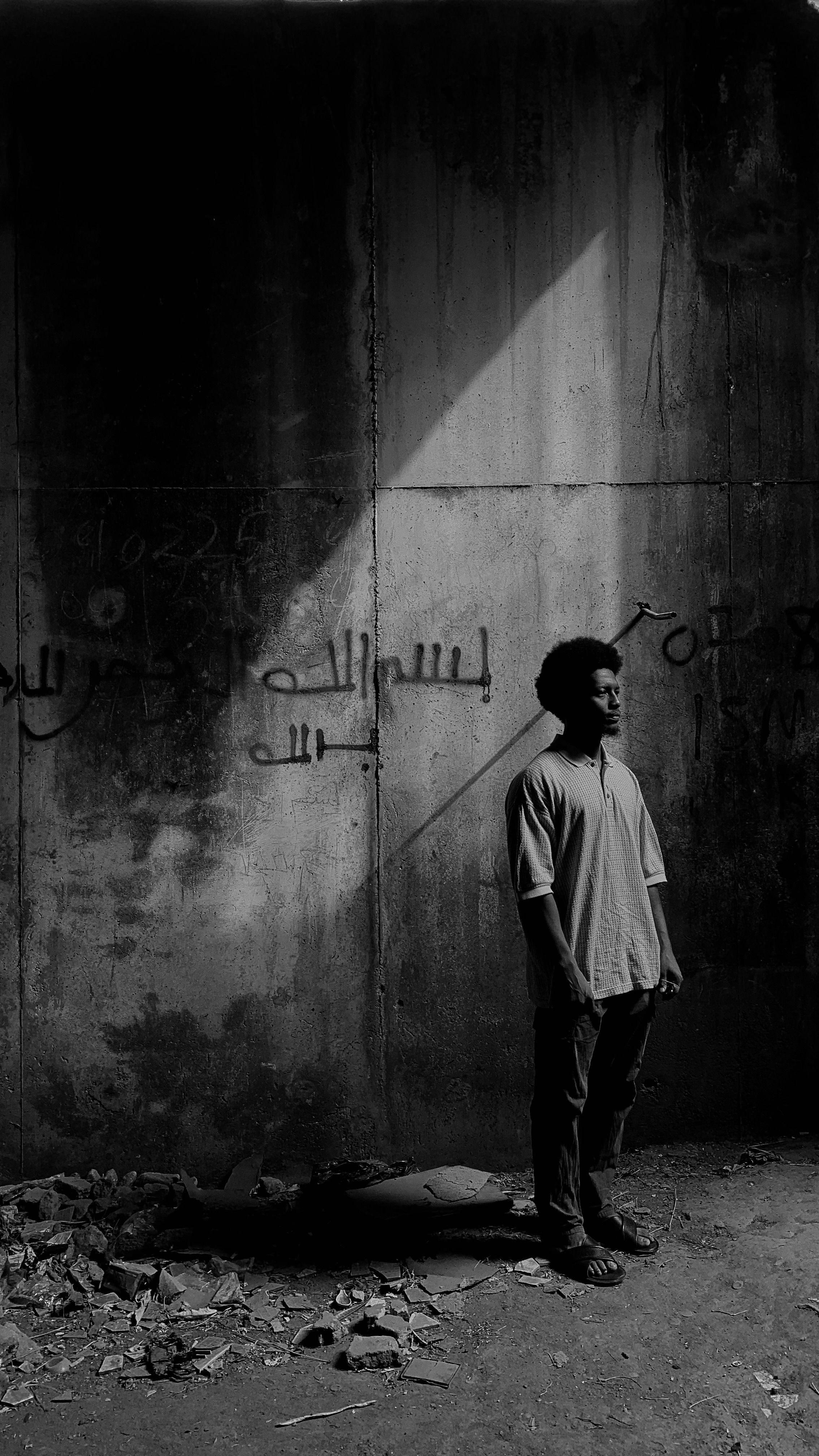 Young man with afro hairstyle stands against graffiti wall, light streaming from above, creating dramatic shadows.