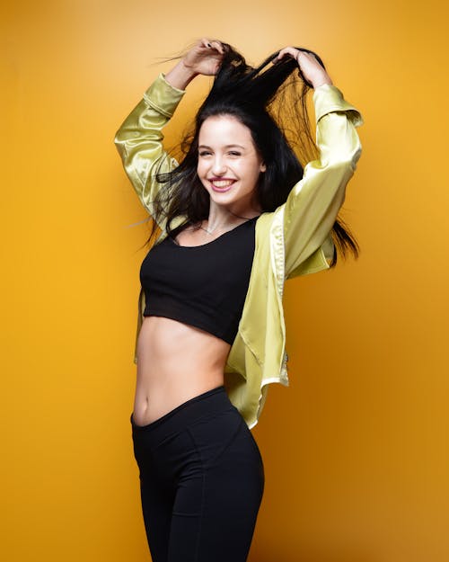 Photo of Smiling Woman in Black Active Wear Standing In Front of Yellow Background While Holding Her Hair