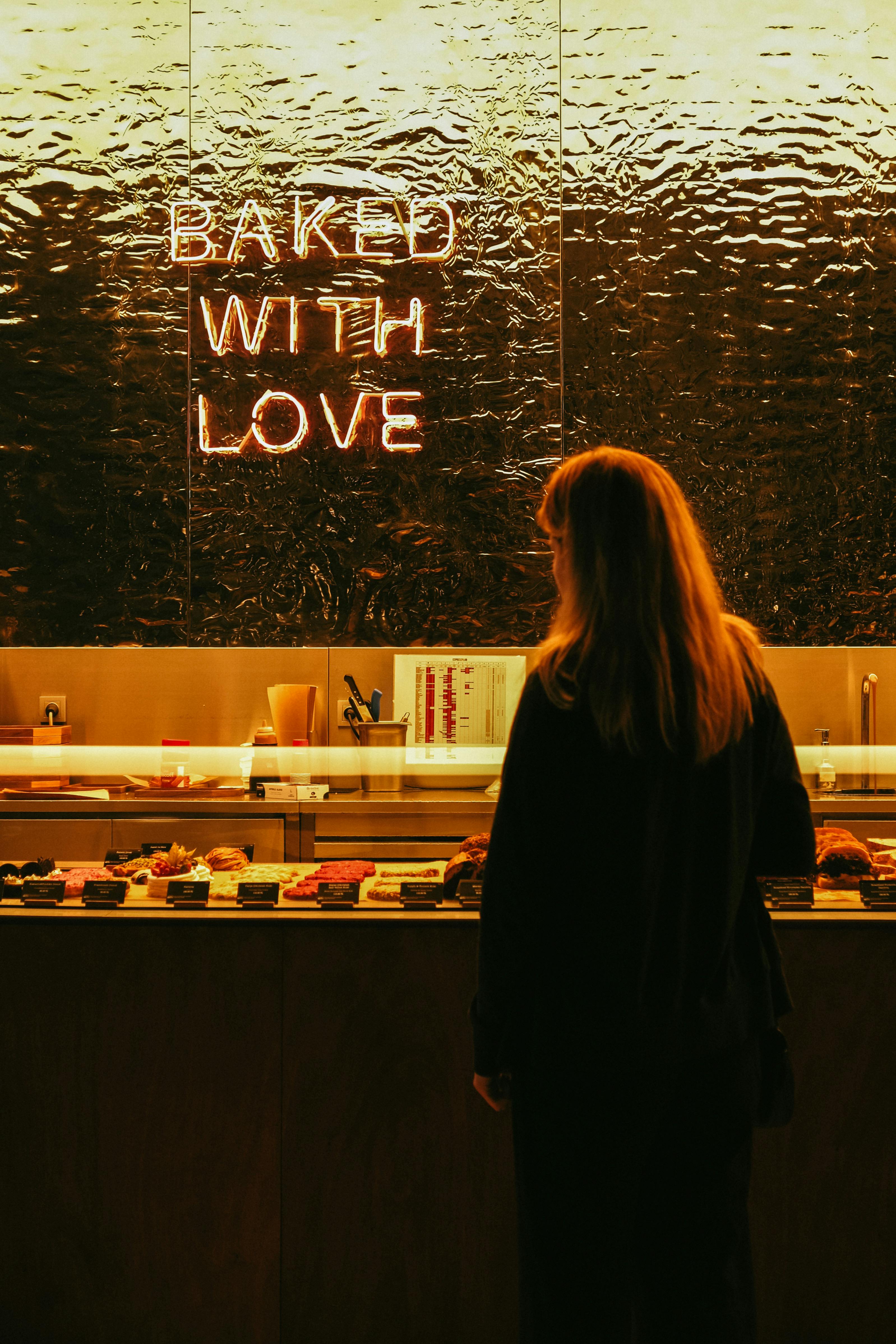 A woman stands before a bakery counter lit by a glowing 'Baked With Love' neon sign, creating a warm and inviting atmosphere.