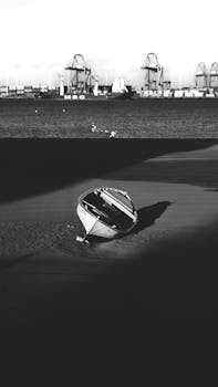 A serene black and white image of a boat on the shore with a distant industrial backdrop.