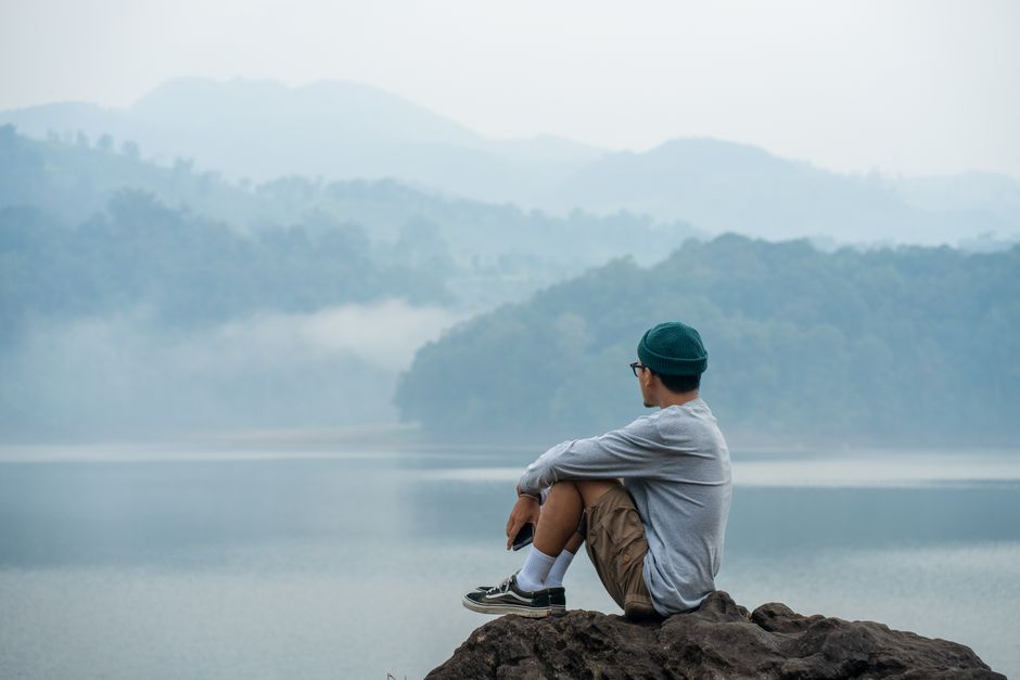 Man relaxing on a boulder by a foggy lake, wearing casual attire, enjoying nature.