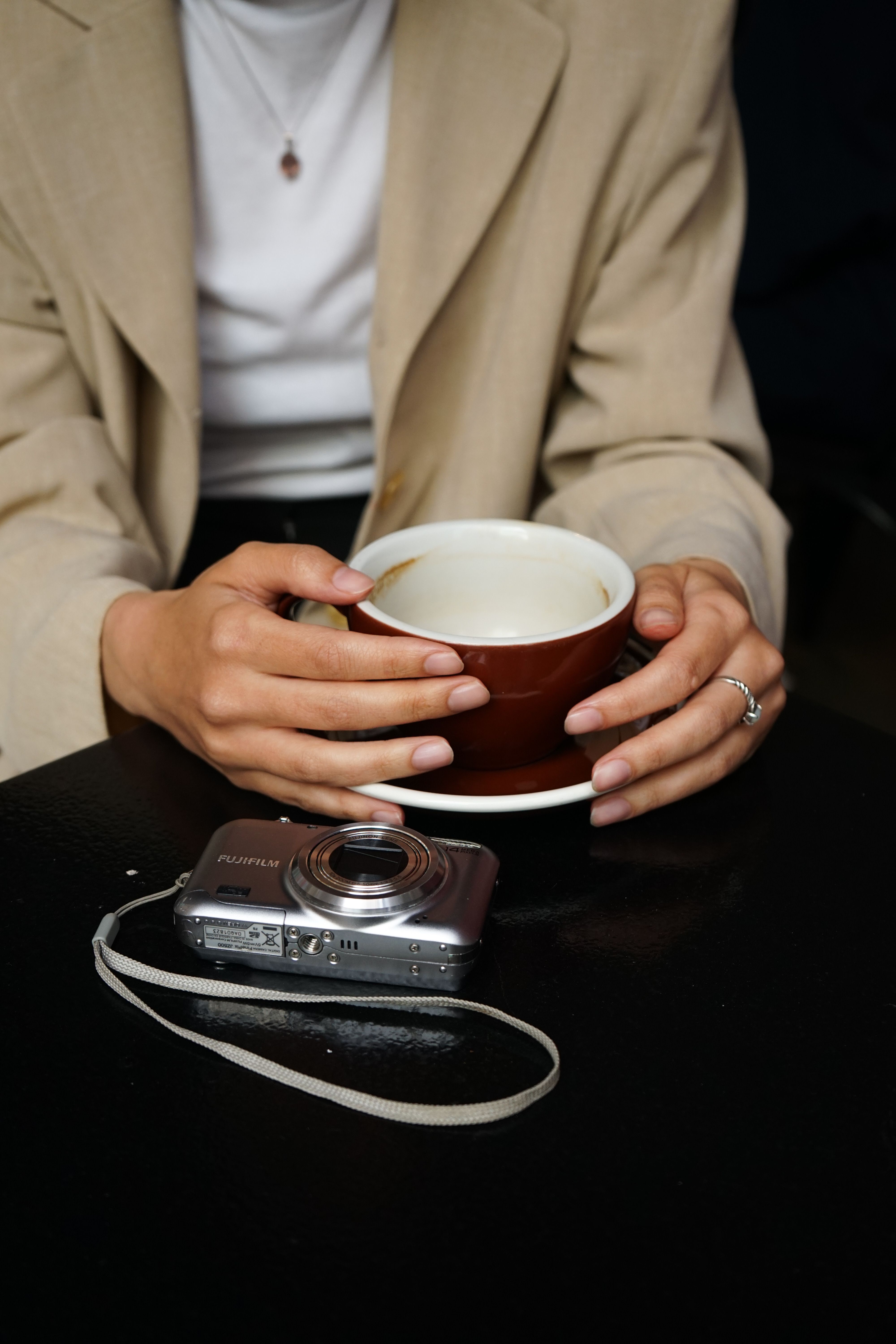 A woman holds a coffee cup with a Fujifilm camera resting on the table, focusing on her hands and rings.. pexels, woman, hold