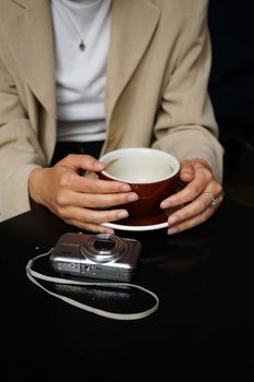 A woman holds a coffee cup with a Fujifilm camera resting on the table, focusing on her hands and rings.. pexels, woman, hold