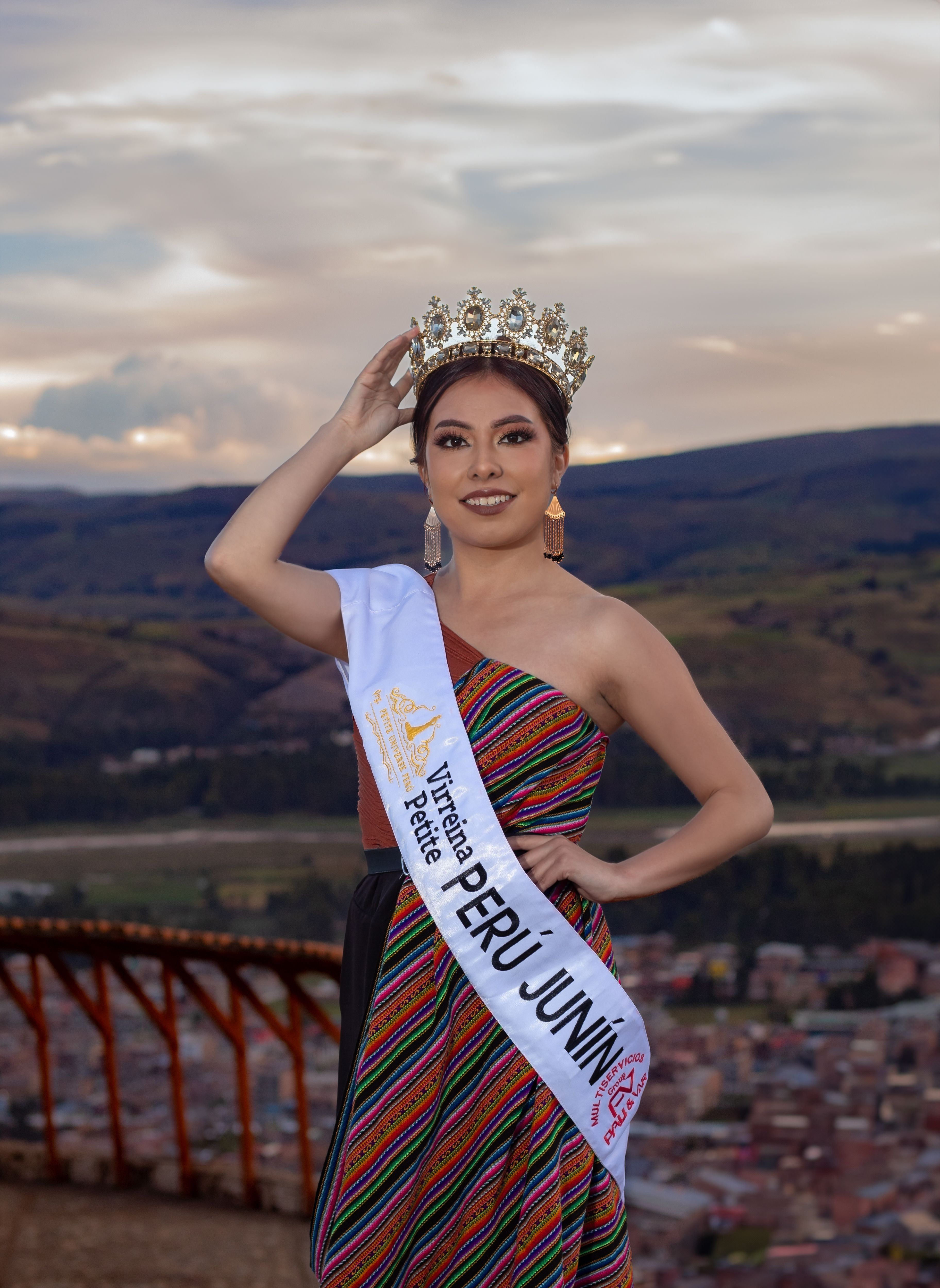 A beauty queen wearing a crown poses in traditional attire with a view of Junín, Perú in the background.