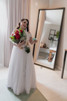 A joyful bride holds a bouquet of red roses while dressed in a beautiful white gown, captured indoors.