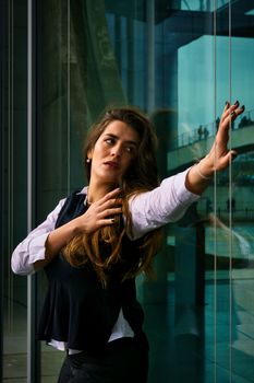 A fashionable woman with long hair poses by a reflective glass window in urban Berlin setting.