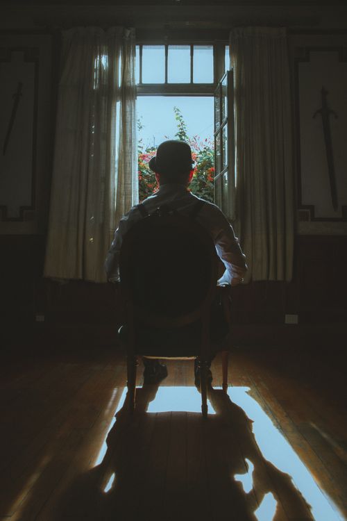 Free A solitary man in a hat sits in a dim room, backlit by sunlight streaming through an open window. Stock Photo