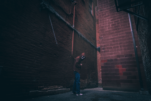 Free A person wearing a mask and hoodie stands in a dimly lit urban alley. Stock Photo