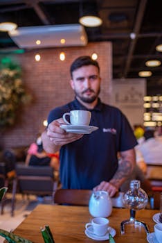 A barista in a modern café extends a cup of coffee towards the camera, creating a welcoming atmosphere.