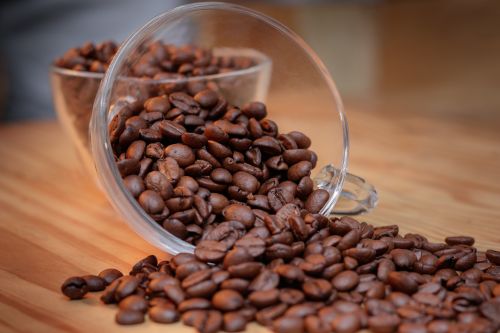 Free A close-up shot of aromatic brown coffee beans spilling from a glass cup on a wooden table. Stock Photo