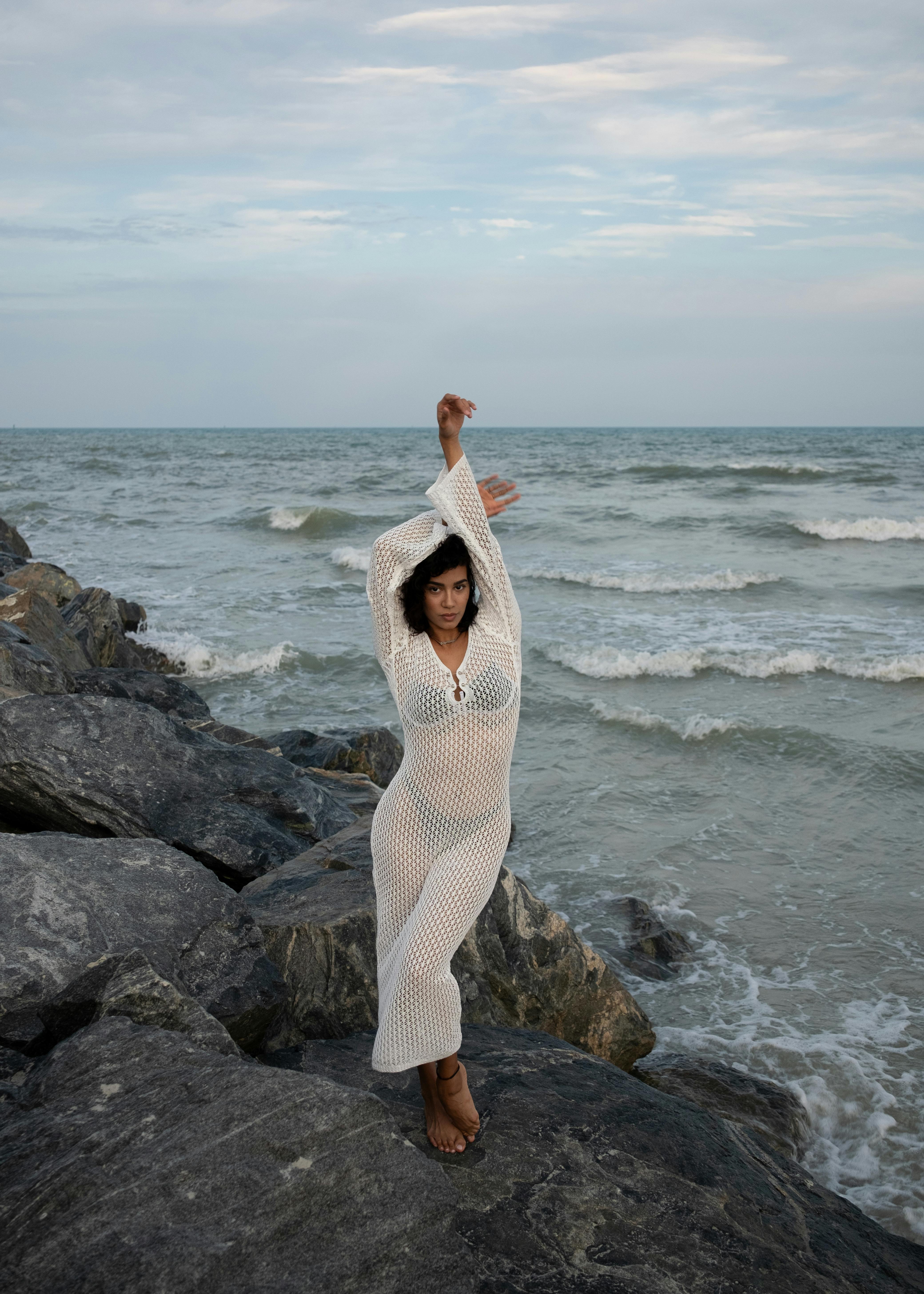 A woman in a white dress posing on rocky shore with ocean waves behind her.