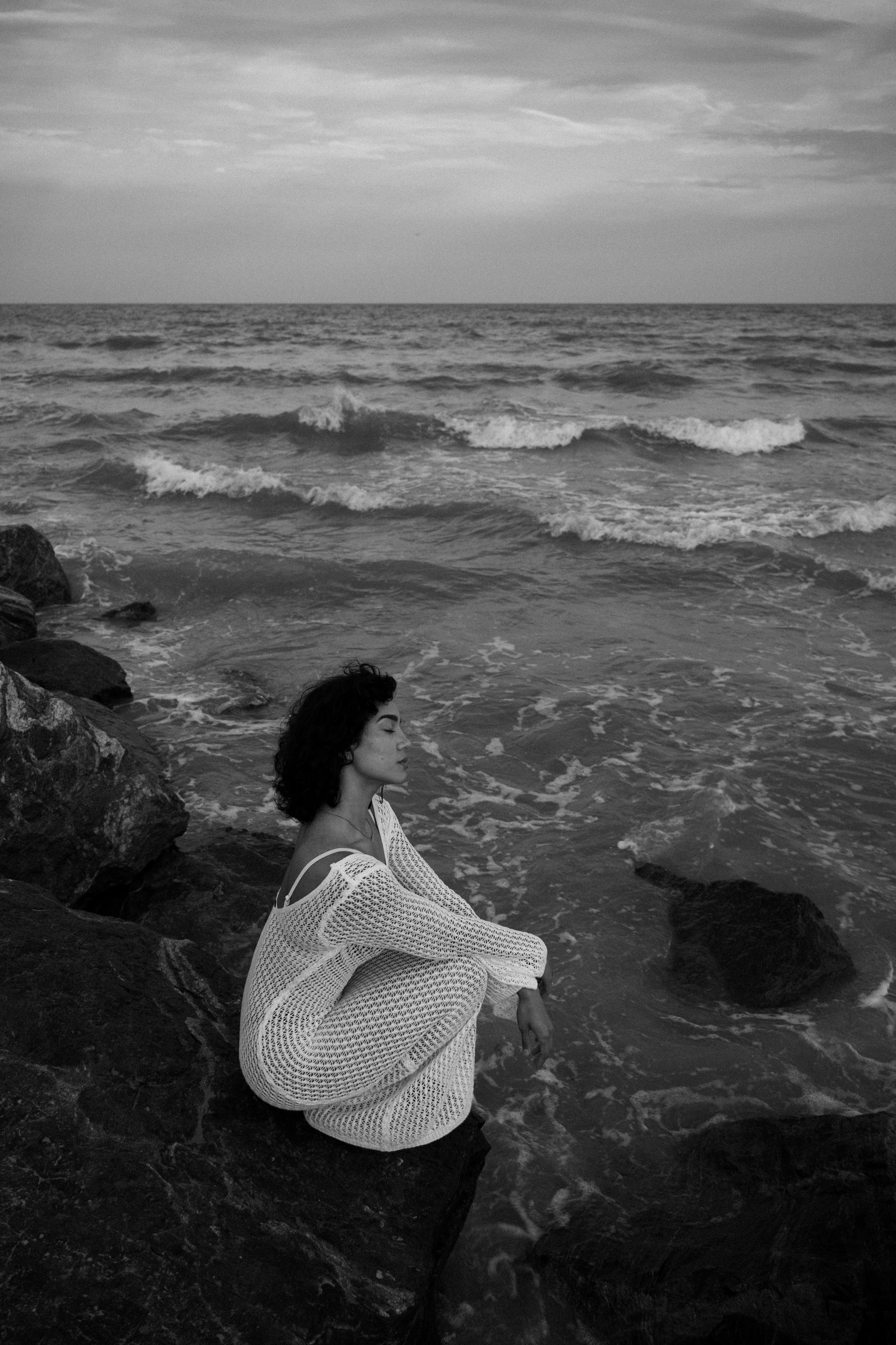 A woman in a white dress poses contemplatively on a rocky shore by the ocean.