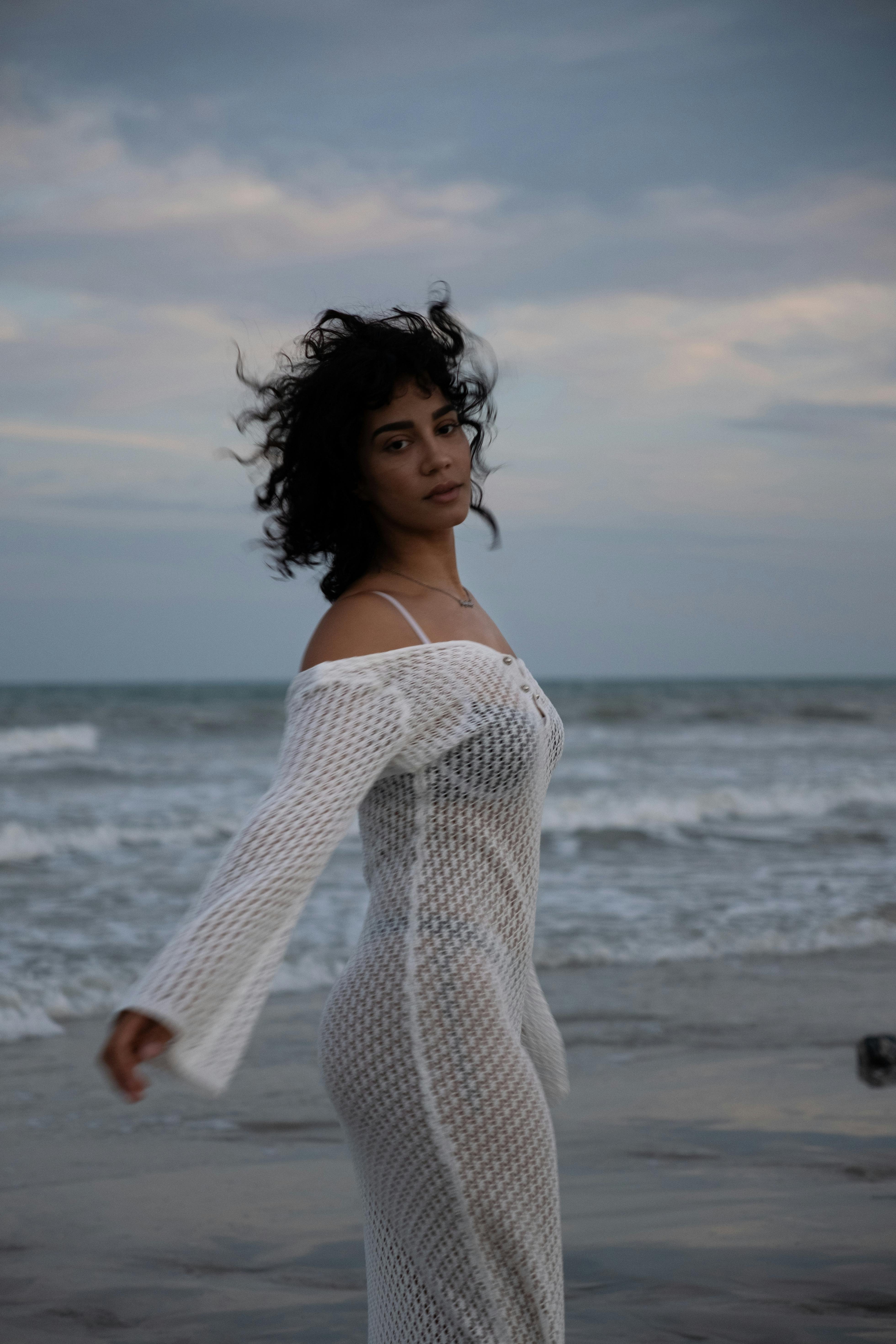 Woman standing by the ocean at sunset, wearing a white dress with wind-blown hair.