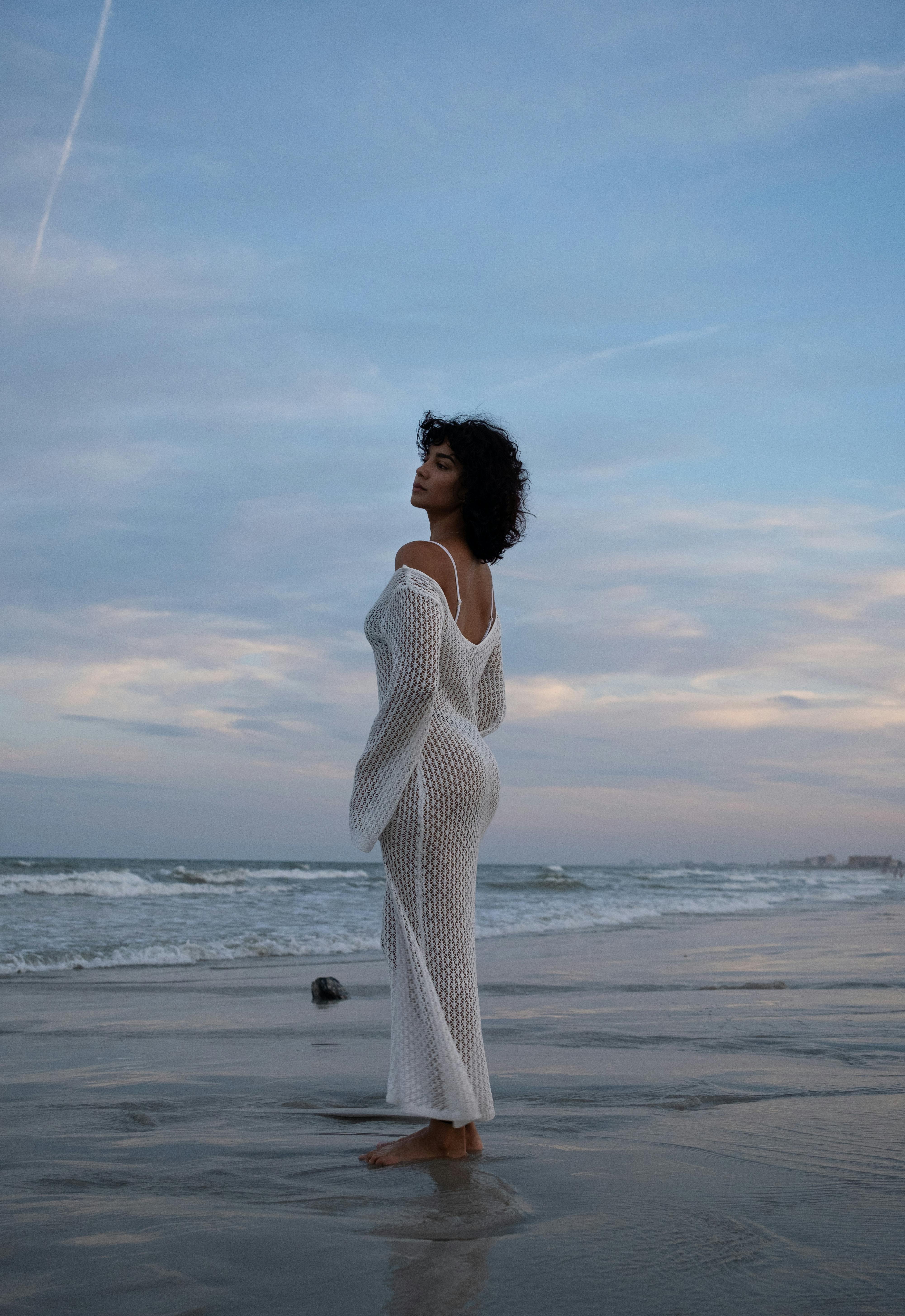 A woman in a white dress poses gracefully on a serene beach with a stunning horizon.