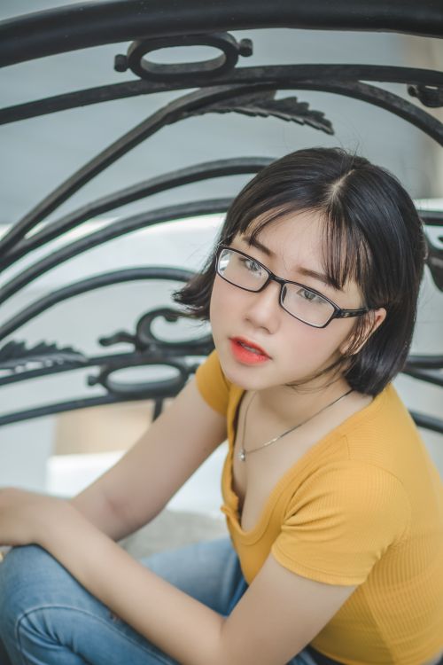 Free Portrait of a young woman in a yellow top and glasses sitting with a thoughtful gaze. Stock Photo