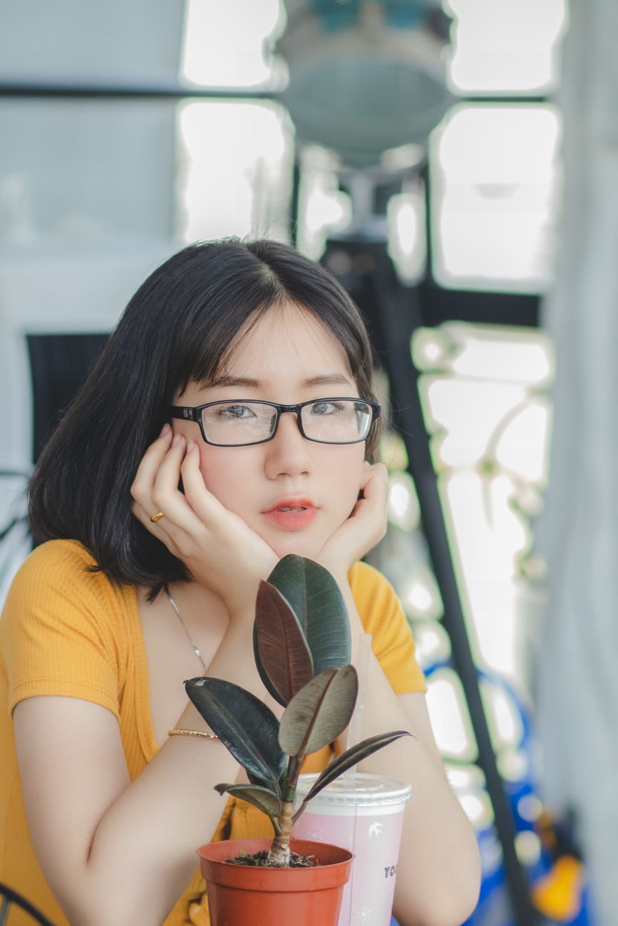 Young woman with glasses, potted plant, and milkshake in a bright indoor space.