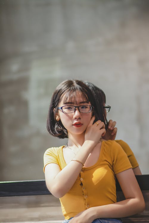 Free Woman in Yellow Scoop Neck Shirt and Black Framed Eyeglasses Standing in Front of Wall Mirror Stock Photo