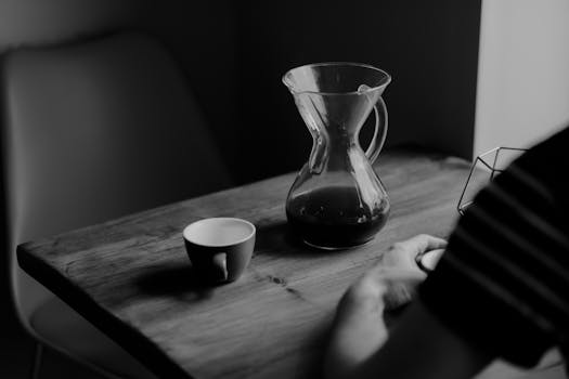 A serene black and white shot of a coffee jug and cup on a wooden table indoors.