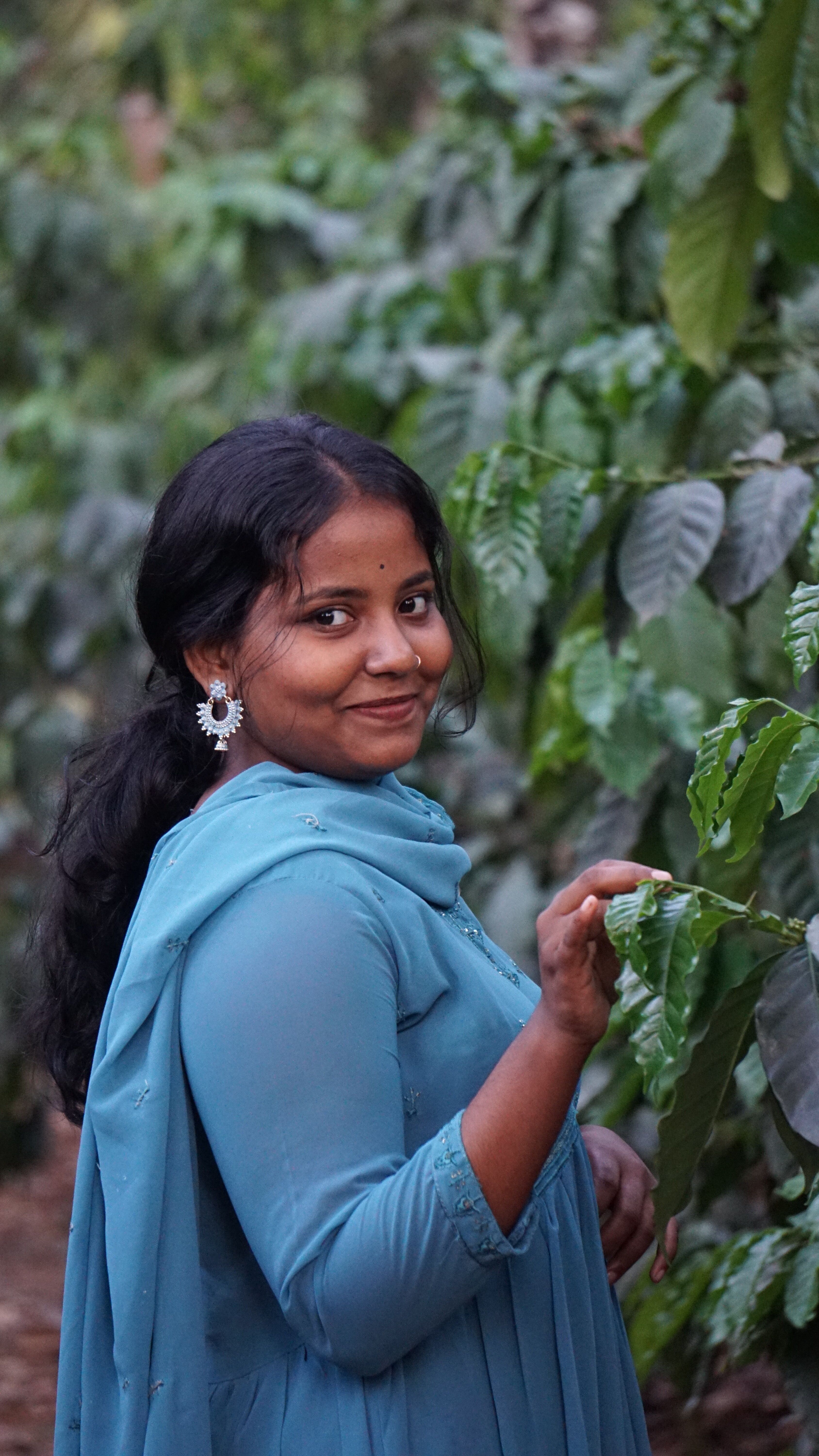 A woman in a blue shawl smiling amidst lush green foliage outdoors.