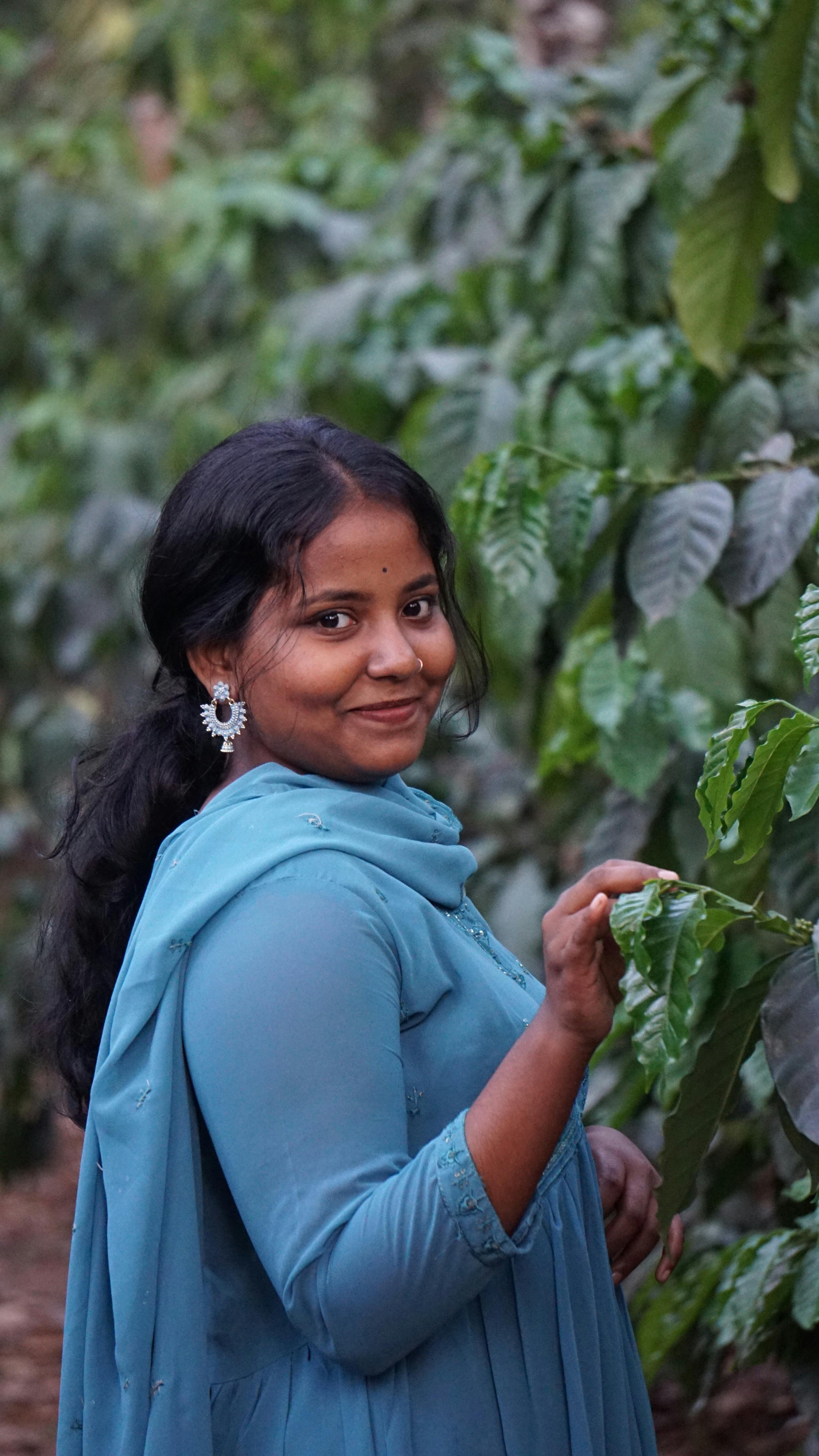 Smiling South Asian woman in blue attire touching lush green plants outdoors.