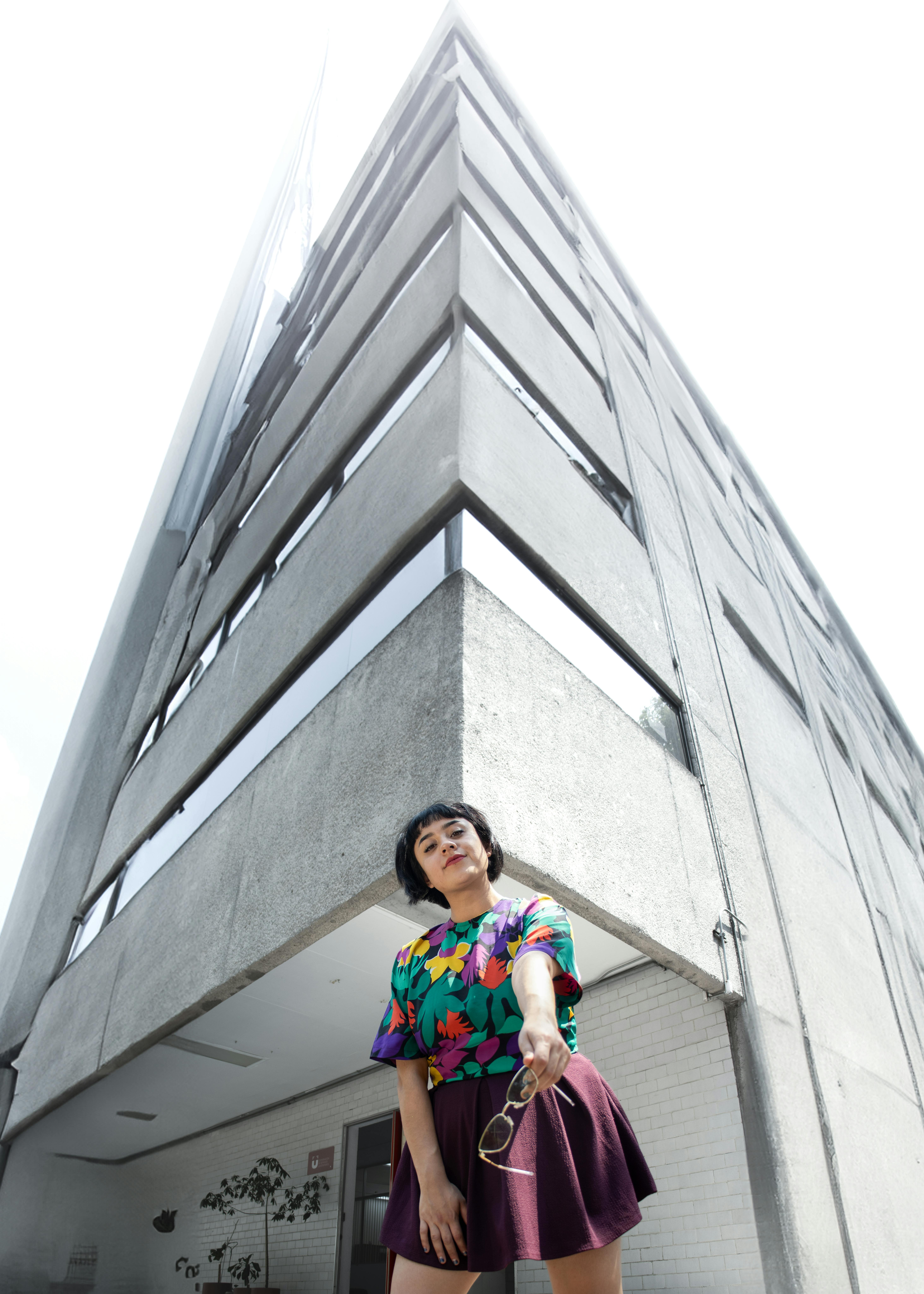Stylish woman in colorful shirt poses under modern triangular building on bright day.