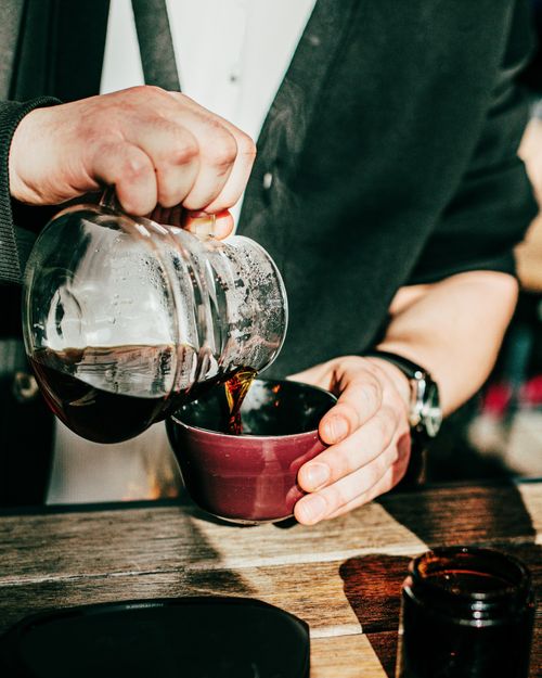 Free Hand pouring coffee into a cup from a glass jug on a rustic wooden table in Baku, Azerbaijan, daytime setting. Stock Photo