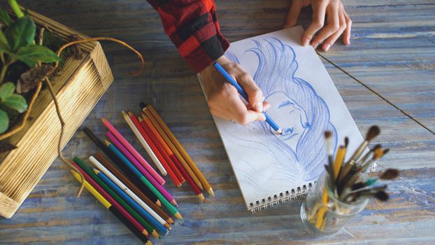 Top view of a person drawing a portrait with color pencils on wooden table.