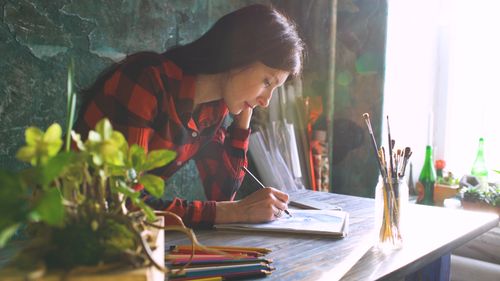 Free A woman in a red plaid shirt sketches at a sunlit table with art supplies nearby. Stock Photo