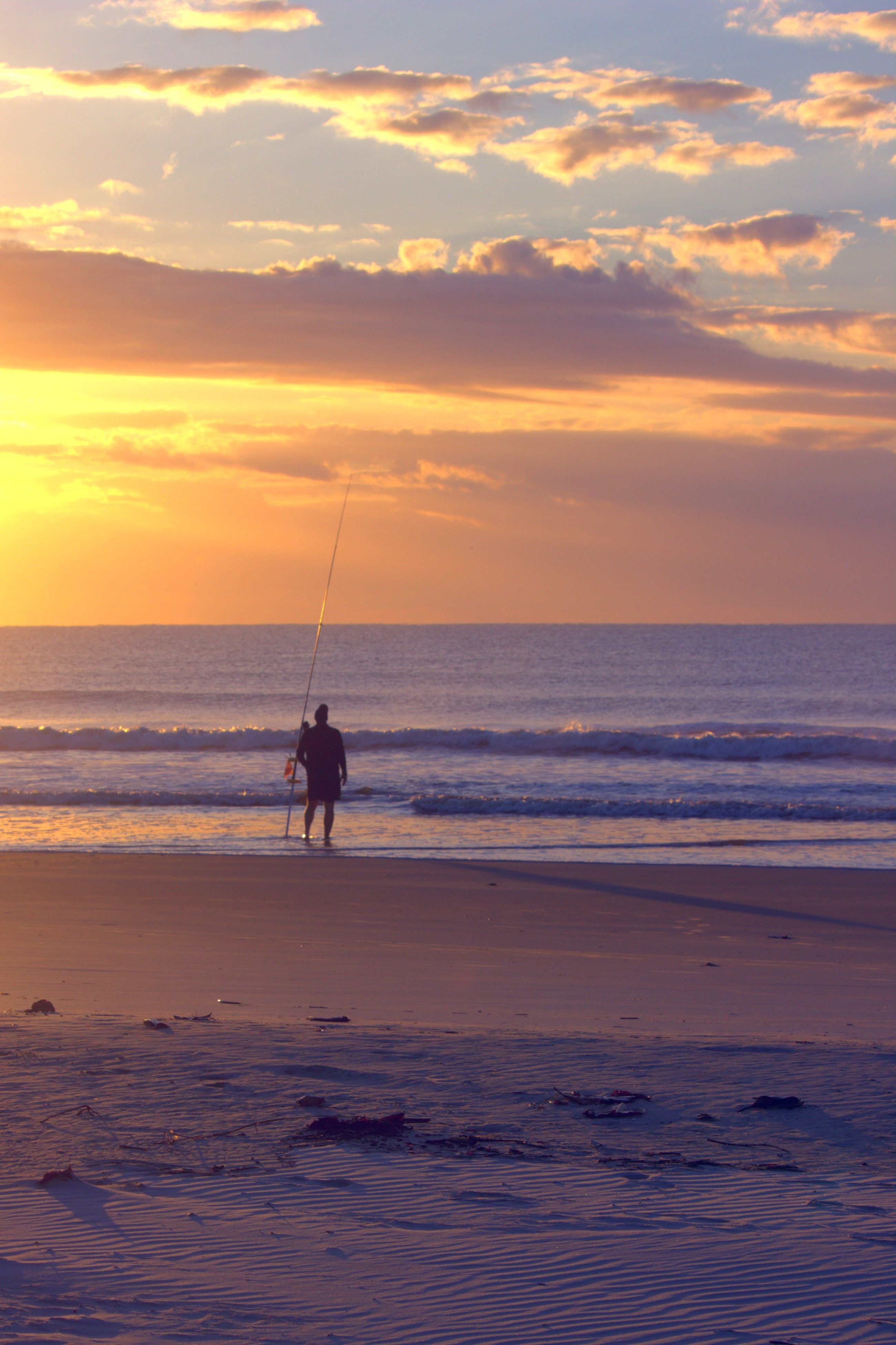 Silhouette of a Fisherman Standing on a Beach at Sunset · Free Stock Photo