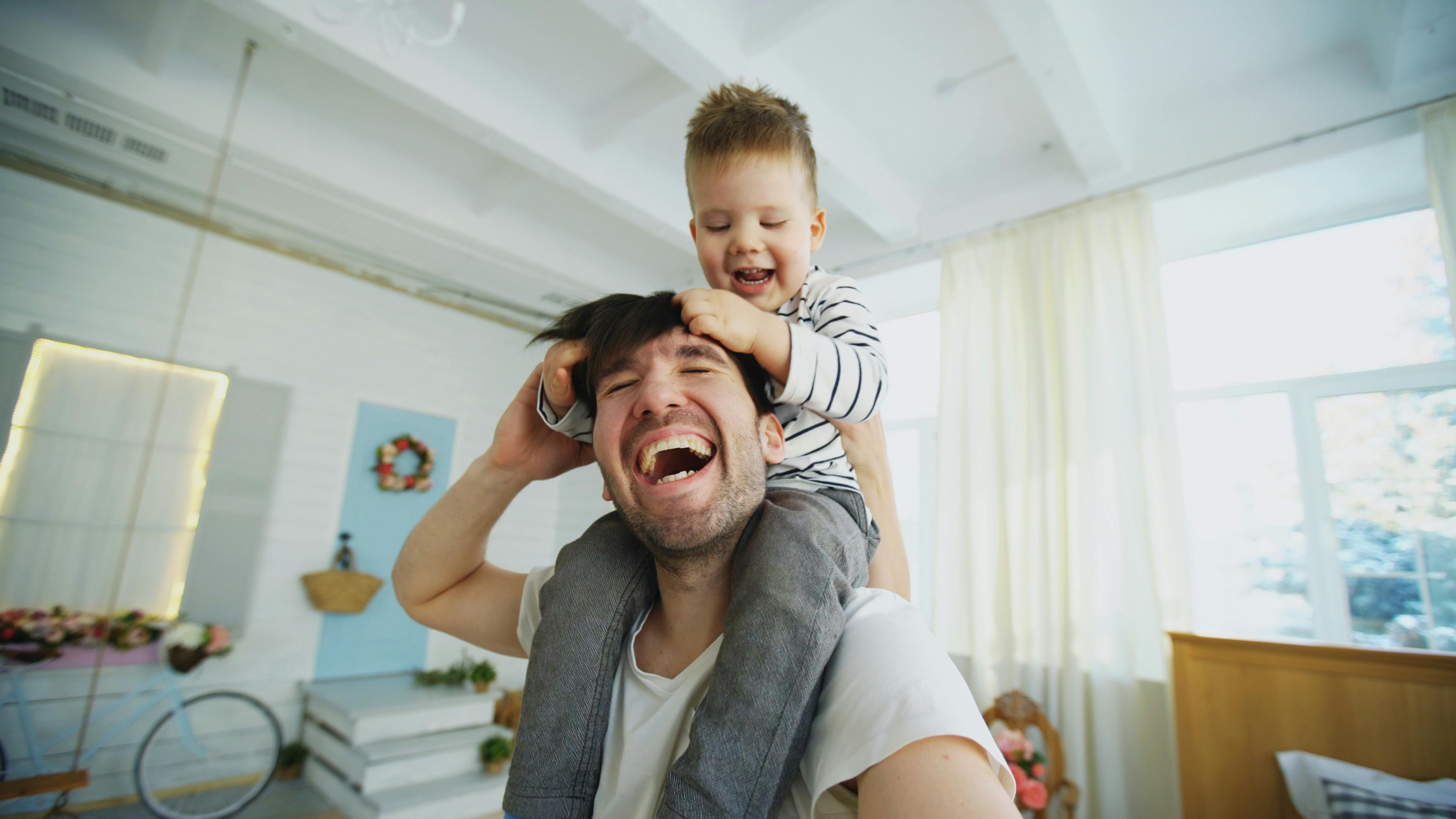 happy father carrying his smiling son on neck and making selfie on smartphone in bedroom