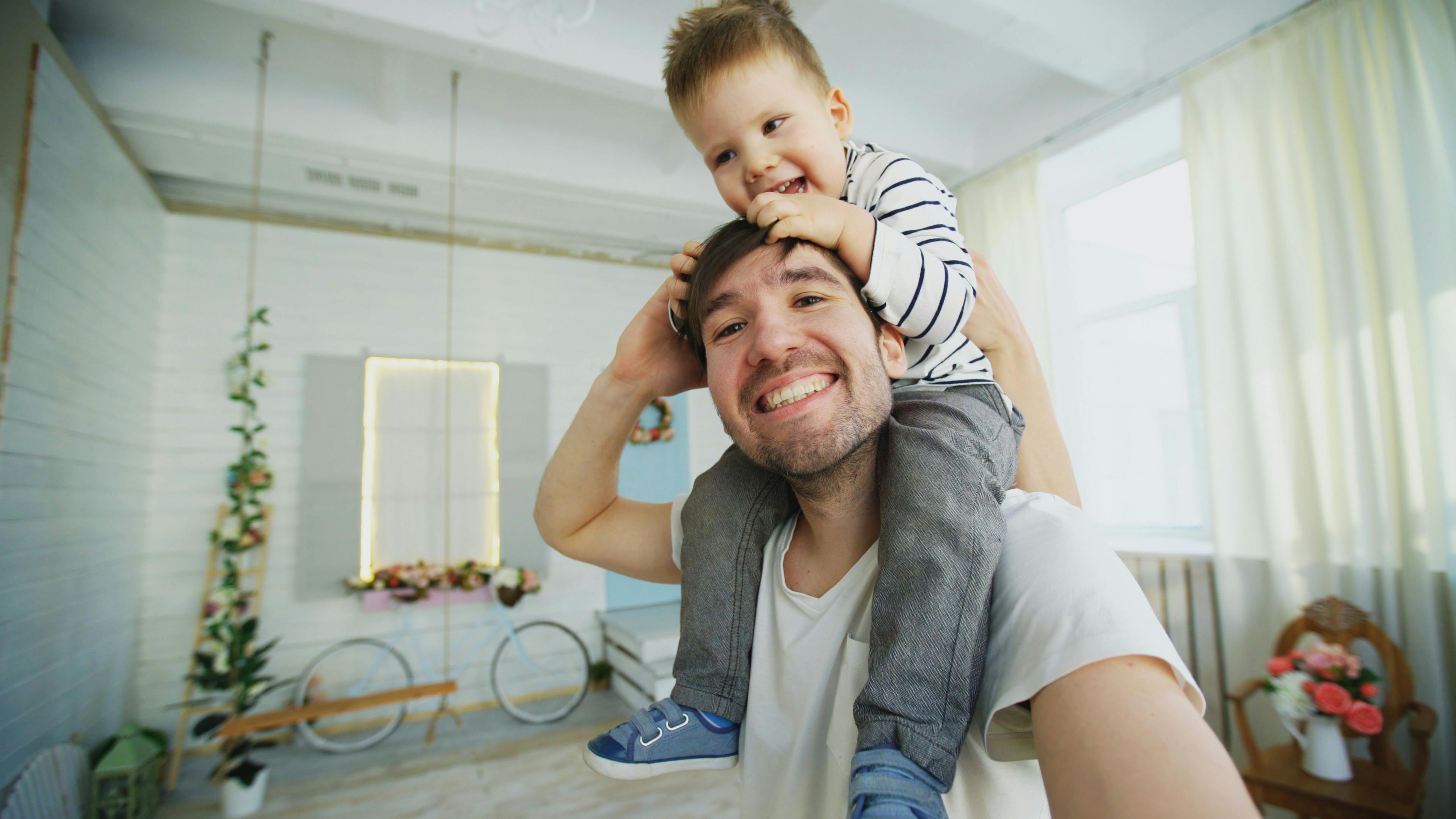 happy father carrying his smiling son on neck and making selfie on smartphone in bedroom
