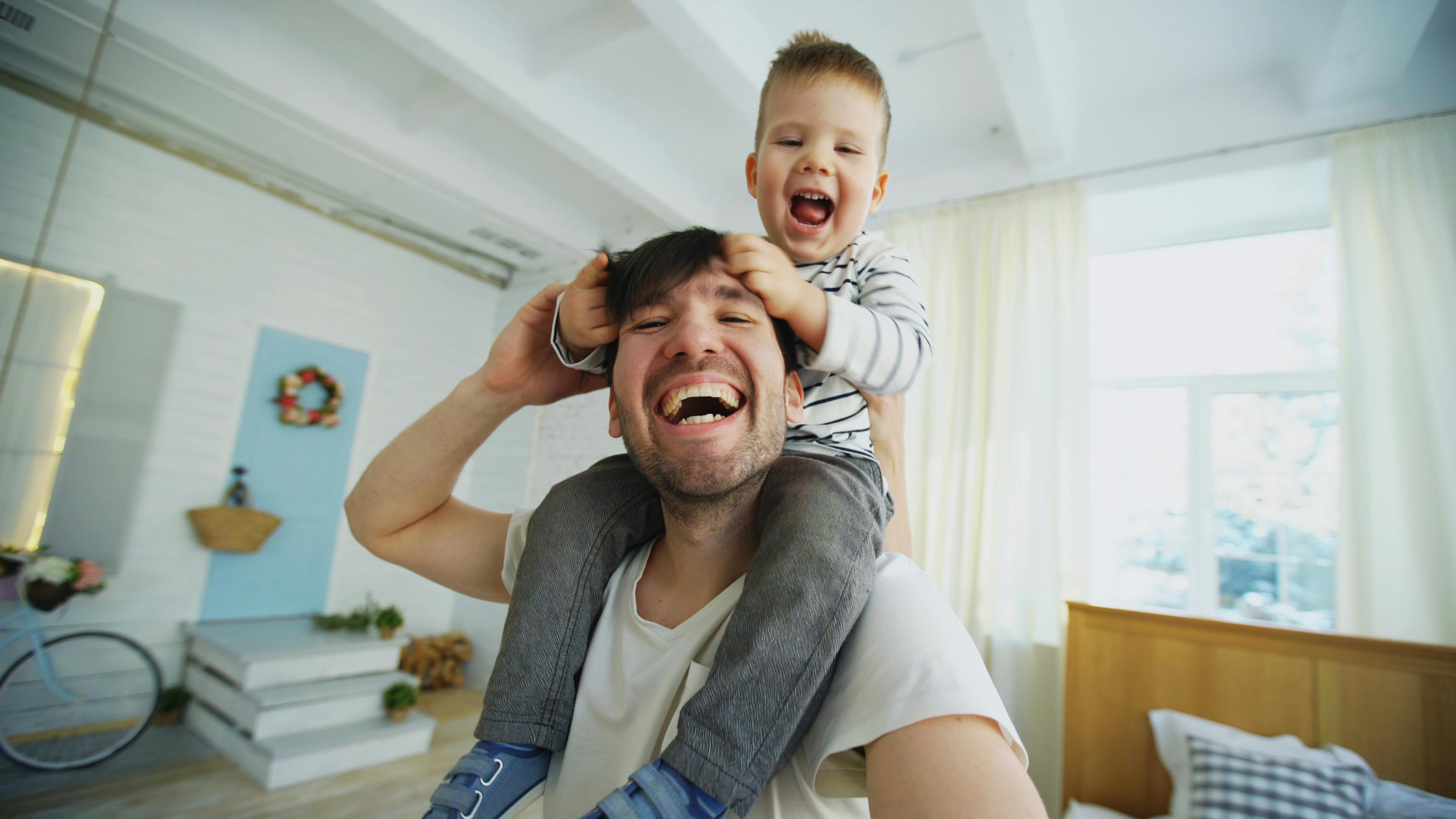 happy father carrying his smiling son on neck and making selfie on smartphone in bedroom