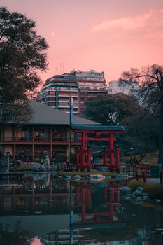 A tranquil Japanese garden with a torii gate reflecting in a pond amidst an urban backdrop at sunset.