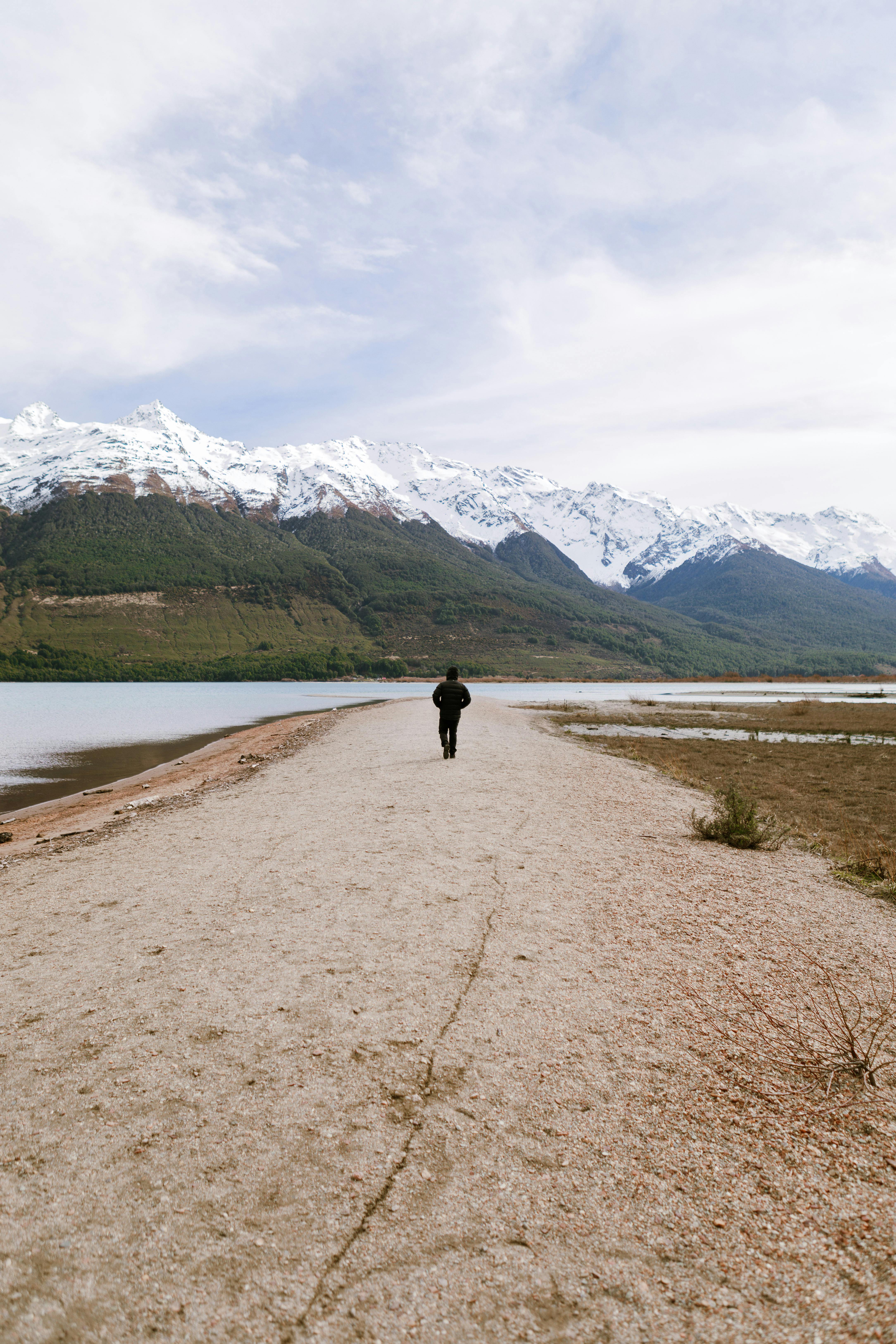 一个孤独的人沿着湖边宁静而风景秀丽的小路散步，周围是白雪皑皑的山脉和郁郁葱葱的绿植