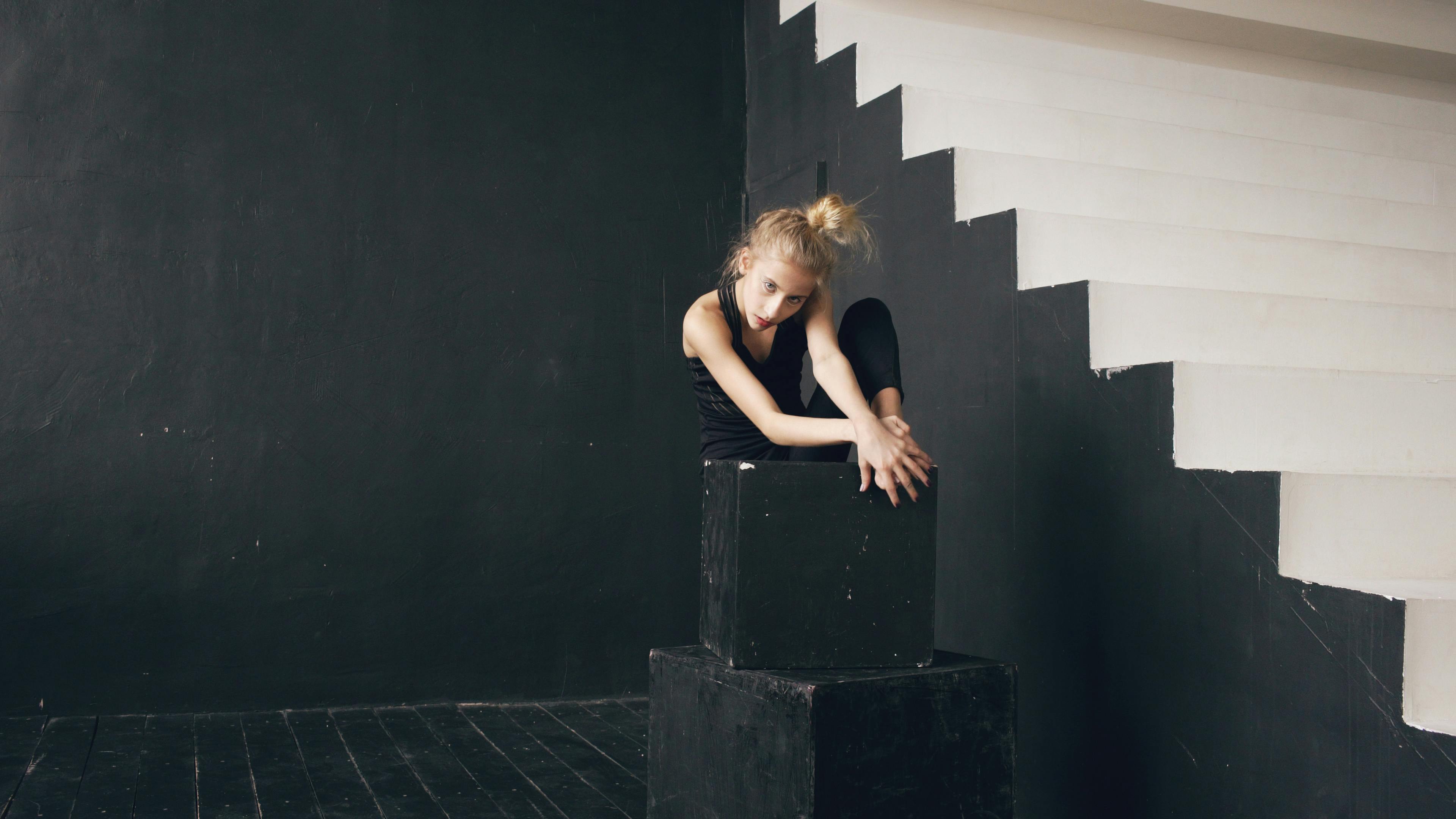 A female dancer posing creatively on black blocks in a minimalist indoor studio setting.