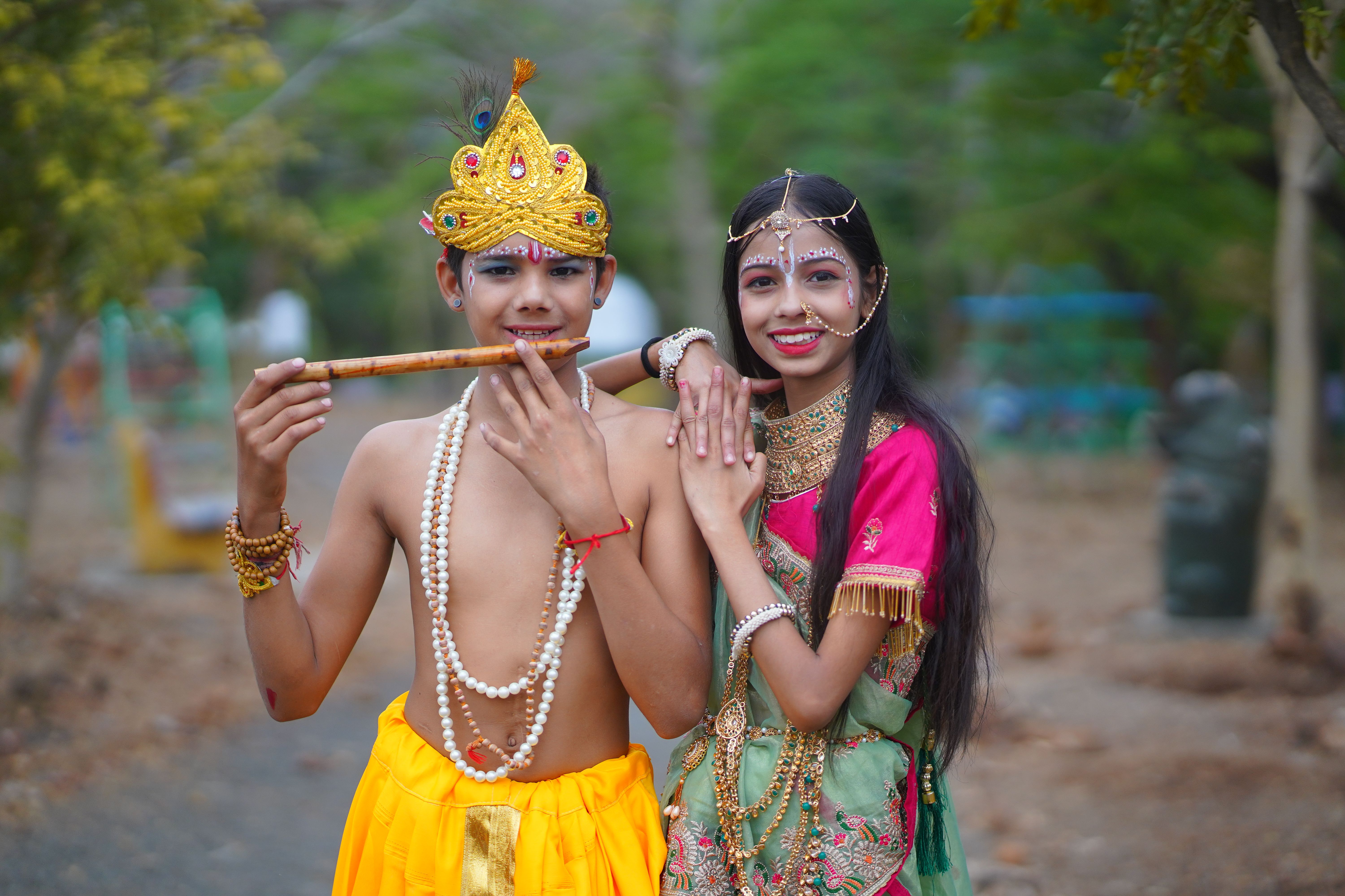 Children dressed as Krishna and Radha in colorful traditional attire, smiling outdoors.