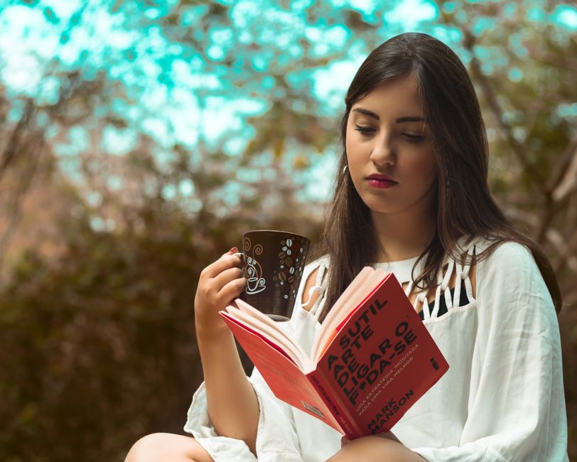 A young woman enjoys reading a book while sipping coffee in a serene outdoor setting.