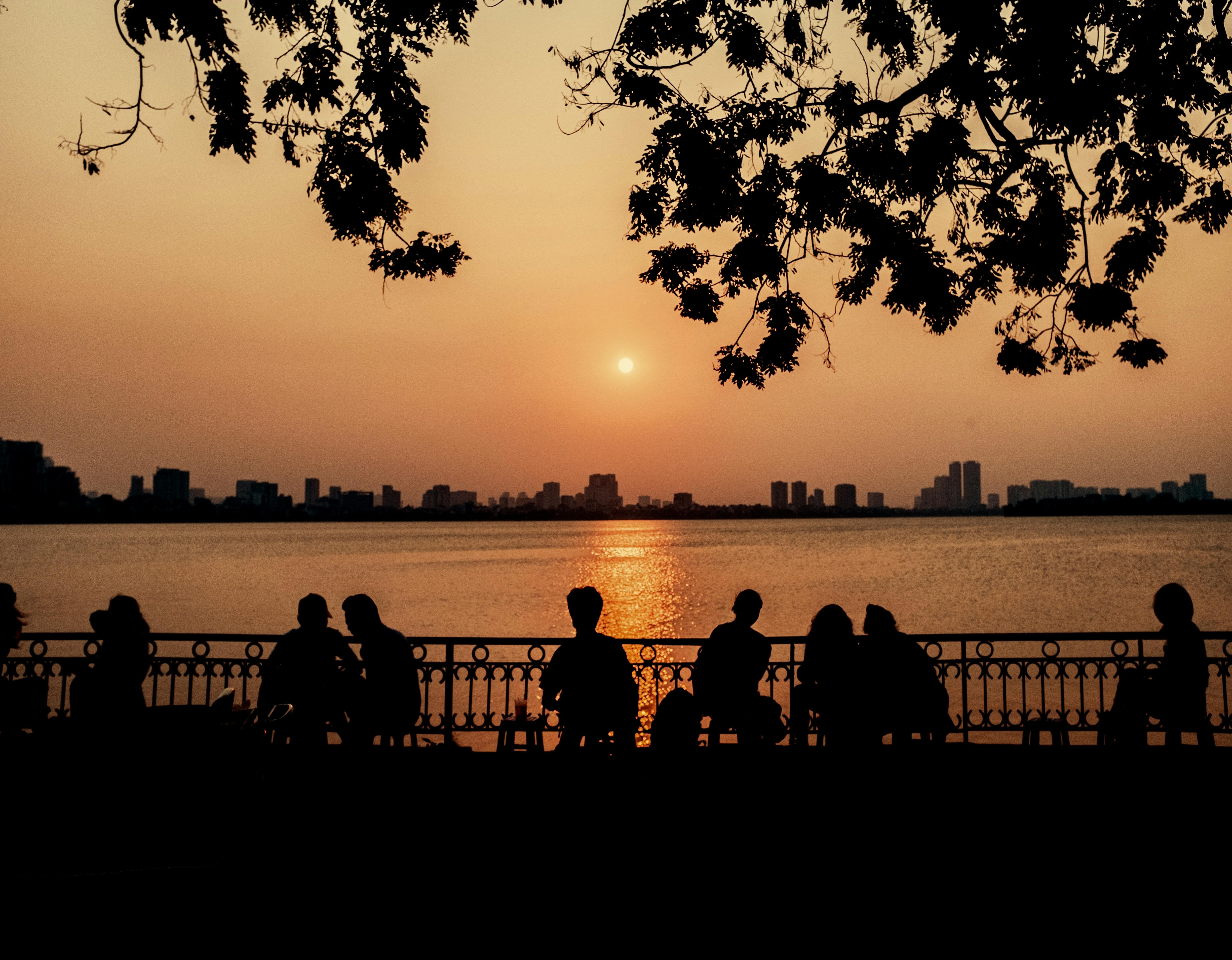 A serene sunset scene with silhouettes of people sitting by the waterfront, cityscape in the background.