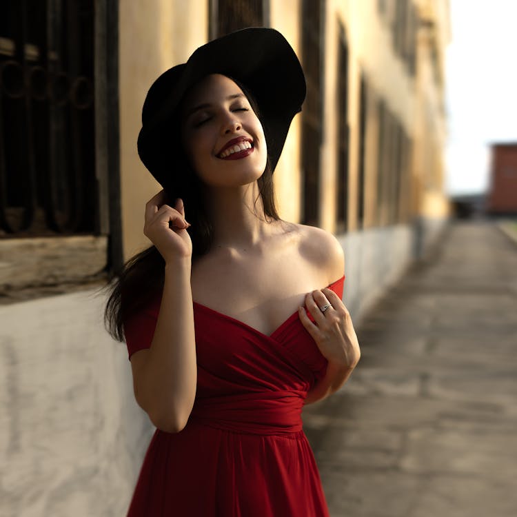 Mujer En Vestido Rojo Con Hombros Descubiertos Sosteniendo Un Sombrero Negro Mientras Mira Hacia Arriba Y Cierra Los Ojos · de stock gratuita