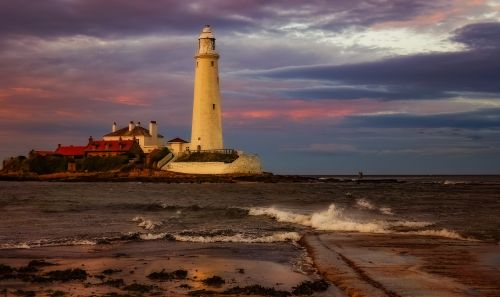 Free Dramatic sunset view of a lighthouse by the ocean, where waves crash against the shore. Stock Photo