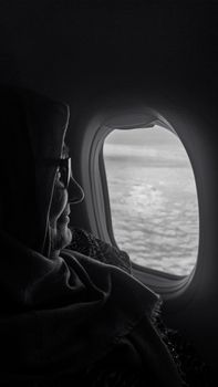 A woman in a hijab looks out an airplane window at the ocean below, capturing a moment of reflection.