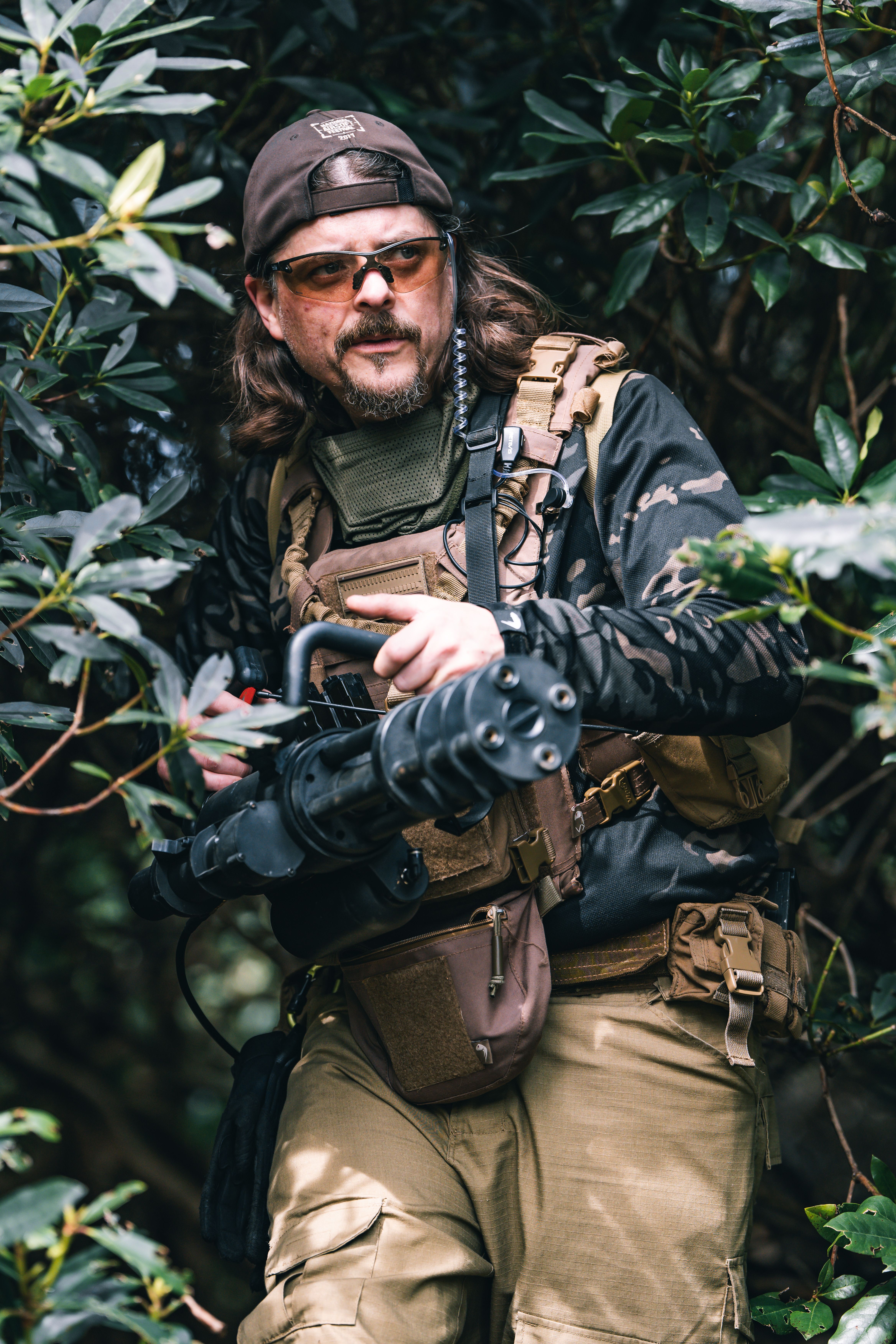 Man with a replica minigun engages in airsoft skirmish in a UK forest setting.
