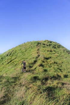 A lone cyclist with mountain bike on a grassy hill; clear blue sky enhances the outdoor adventure vibe.
