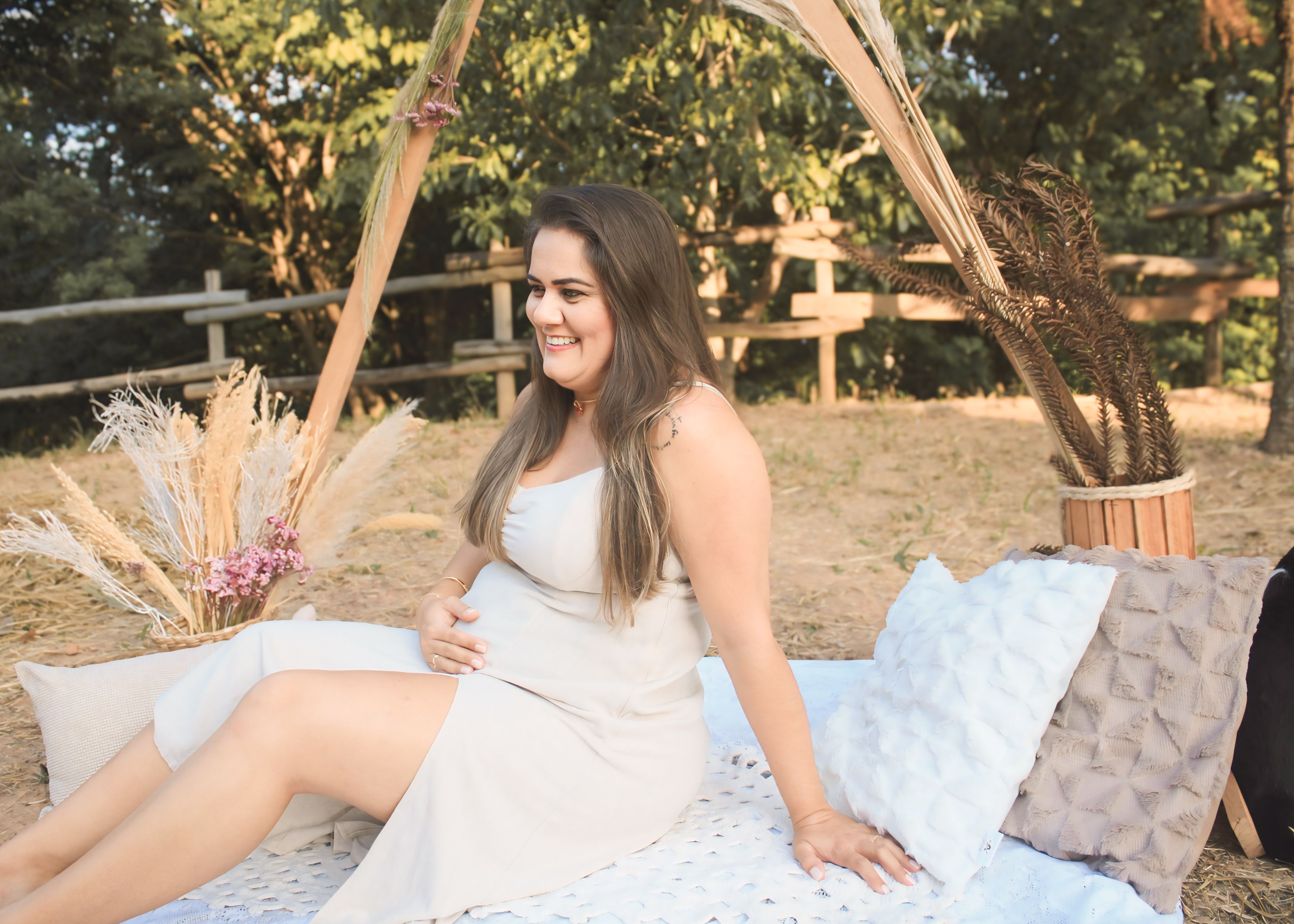 Pregnant woman smiling outdoors, sitting on a blanket with natural decor.