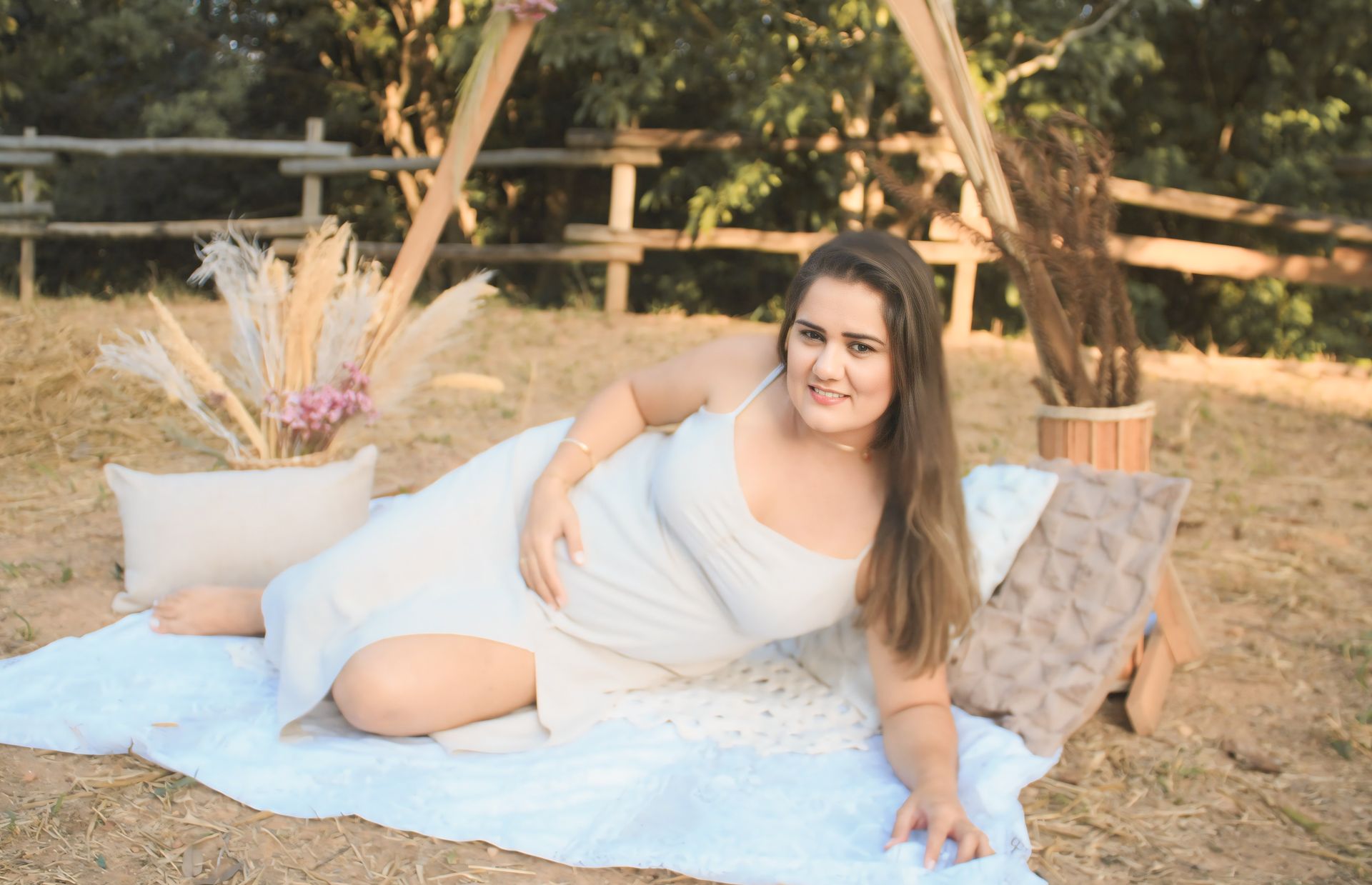 A pregnant woman lies comfortably on a blanket outdoors, surrounded by natural decor.