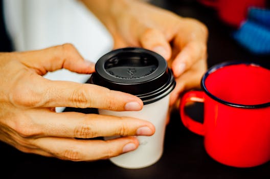 A close-up image of hands holding a disposable coffee cup with a plastic lid next to a red mug.