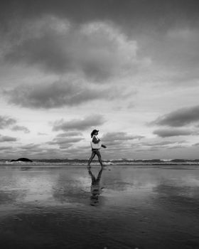 A woman strolls along La Lucila del Mar beach under dramatic clouds, captured in black and white.